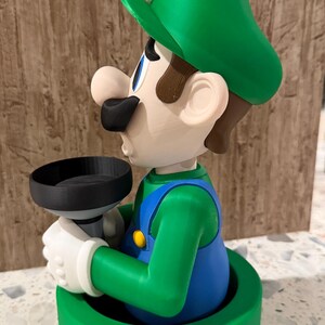 May include: A 3D-printed figurine of Luigi from the Super Mario Bros. series. The figure is in a green pipe, wearing his signature green hat, blue overalls, and white gloves. He is holding a black object.