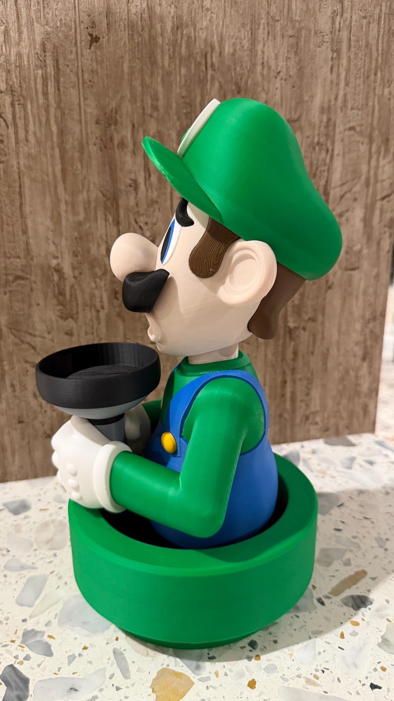 May include: A 3D-printed figurine of Luigi from the Super Mario Bros. series. The figure is in a green pipe, wearing his signature green hat, blue overalls, and white gloves. He is holding a black object.