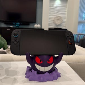 Pode incluir: Uma consola Nintendo Switch preta repousa sobre um suporte roxo com o tema Gengar. O suporte apresenta um Gengar sorridente com olhos vermelhos e uma base de nuvem roxa. A consola tem comandos azuis e laranja.