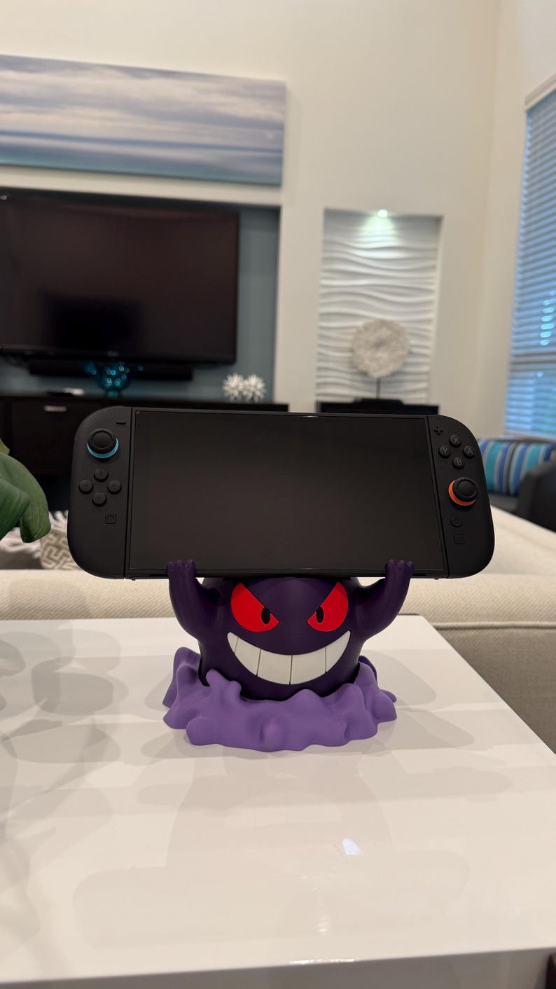 Pode incluir: Uma consola Nintendo Switch preta repousa sobre um suporte roxo com o tema Gengar. O suporte apresenta um Gengar sorridente com olhos vermelhos e uma base de nuvem roxa. A consola tem comandos azuis e laranja.