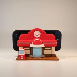 May include: A red and white 3D-printed model of a building with a Pok&eacute; Ball logo, a small red phone, and a black game console in the background. The model is on a brown base. The building has a red roof and white accents.