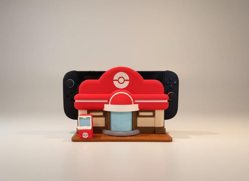 May include: A red and white 3D-printed model of a building with a Pok&eacute; Ball logo, a small red phone, and a black game console in the background. The model is on a brown base. The building has a red roof and white accents.