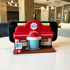 May include: A red and white decorative structure resembling a building, with a blue door and a small red kiosk. A black Nintendo Switch console is placed behind the structure. The structure is on a brown base, and the background is a white table.