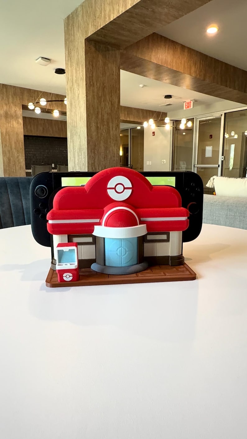 May include: A red and white decorative structure resembling a building, with a blue door and a small red kiosk. A black Nintendo Switch console is placed behind the structure. The structure is on a brown base, and the background is a white table.