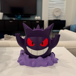 Pode incluir: Uma figura Gengar roxa com olhos vermelhos e um sorriso largo. A figura tem uma base em forma de nuvem e est&aacute; posada com os bra&ccedil;os levantados. A figura est&aacute; sobre uma superf&iacute;cie branca.