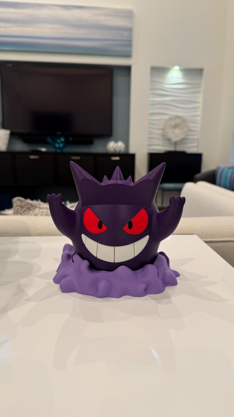 Pode incluir: Uma figura Gengar roxa com olhos vermelhos e um sorriso largo. A figura tem uma base em forma de nuvem e est&aacute; posada com os bra&ccedil;os levantados. A figura est&aacute; sobre uma superf&iacute;cie branca.