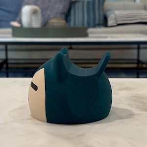 May include: A teal-coloured, cartoon-style figurine with a cream-coloured face and black eyes. The figurine has a rounded body with two small ears and a small tail. The object is sitting on a white marble surface.