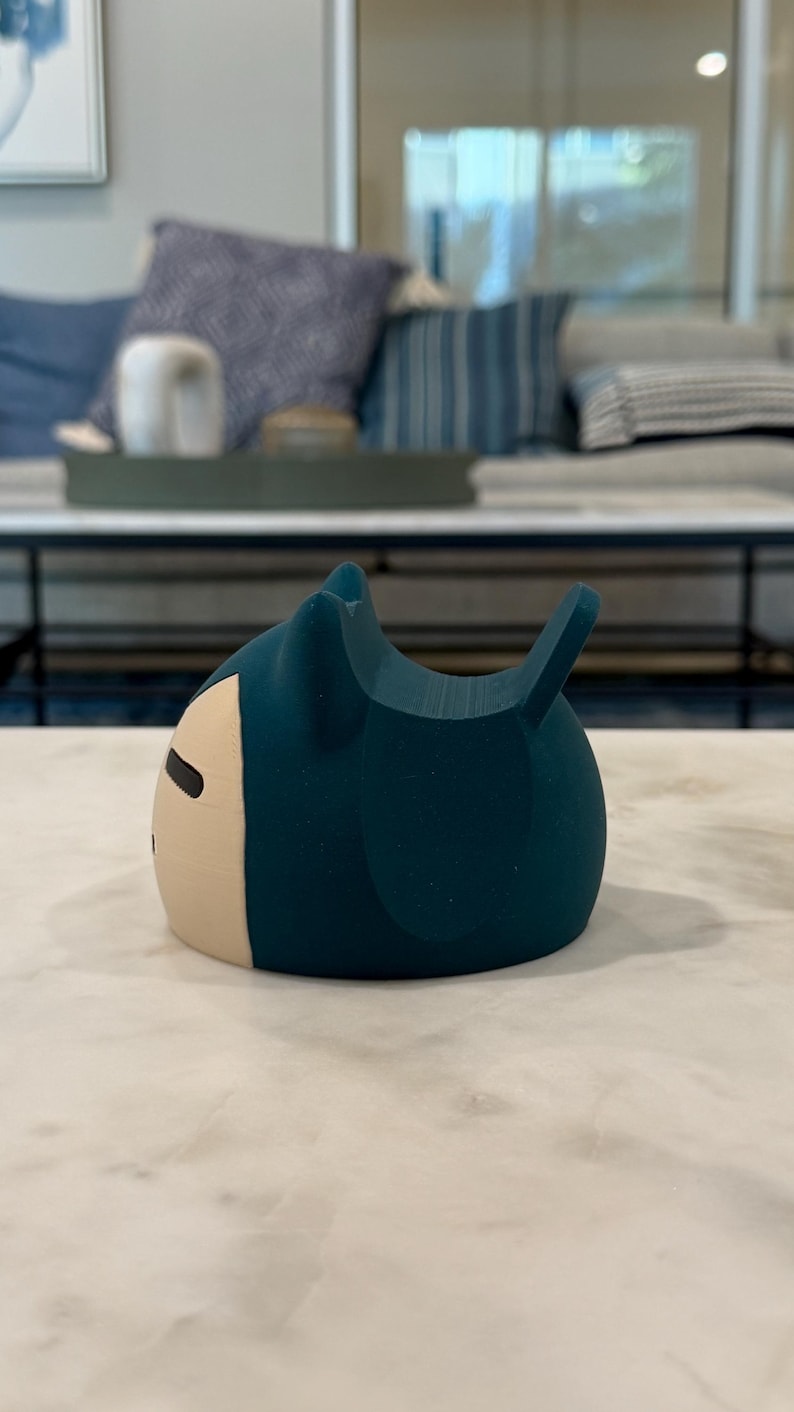 May include: A teal-coloured, cartoon-style figurine with a cream-coloured face and black eyes. The figurine has a rounded body with two small ears and a small tail. The object is sitting on a white marble surface.