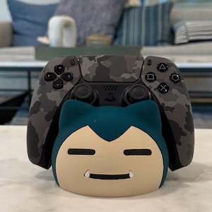 May include: A grey camouflage-patterned video game controller sits on a cream-coloured stand shaped like a cartoon character with a teal head and a beige face. The controller has black buttons and a black base.