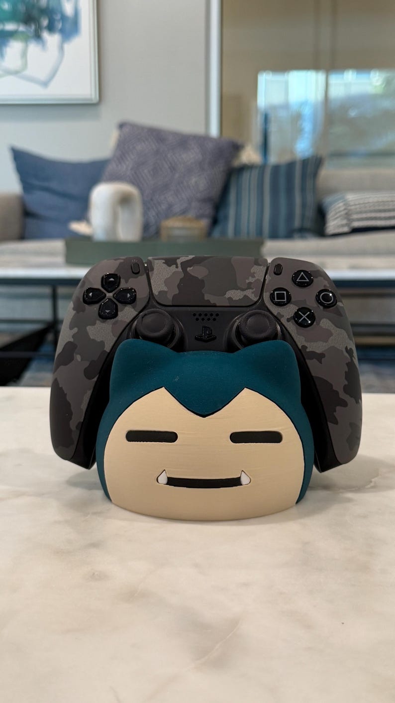 May include: A grey camouflage-patterned video game controller sits on a cream-coloured stand shaped like a cartoon character with a teal head and a beige face. The controller has black buttons and a black base.