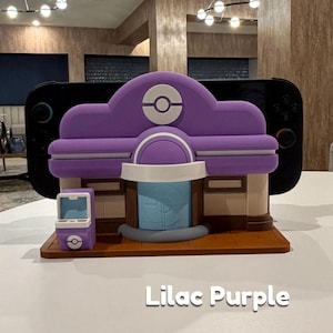 May include: A lilac purple 3D-printed model of a building with a rounded roof, a blue door, and a small matching kiosk. The model has a Pok&eacute;mon logo on the roof. The text "Lilac Purple" is below the model.