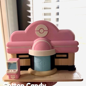 May include: A pink and beige 3D-printed model of a building with a blue door and a small pink vending machine. The building has a rounded roof with a white and pink logo. The text "Cotton Candy" is at the bottom.
