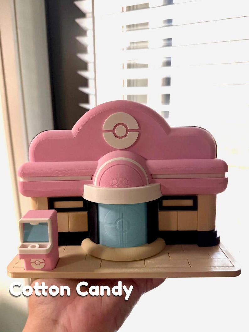 May include: A pink and beige 3D-printed model of a building with a blue door and a small pink vending machine. The building has a rounded roof with a white and pink logo. The text "Cotton Candy" is at the bottom.