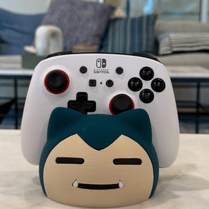 May include: A white Nintendo Switch controller with black buttons and red accents sits on a teal and tan Snorlax-shaped stand. The controller has the Nintendo Switch logo. The stand is a decorative item for video game accessories.