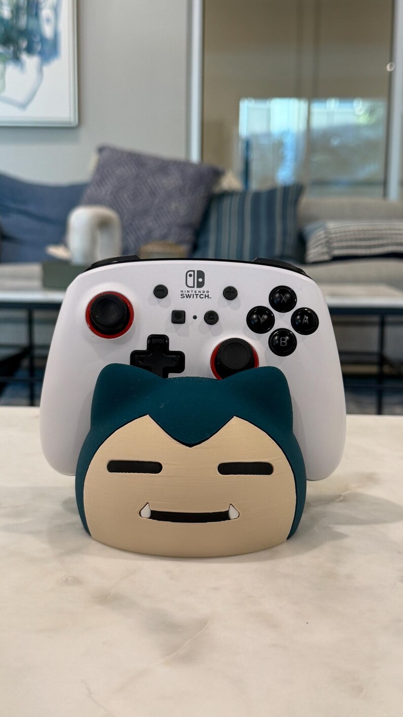 May include: A white Nintendo Switch controller with black buttons and red accents sits on a teal and tan Snorlax-shaped stand. The controller has the Nintendo Switch logo. The stand is a decorative item for video game accessories.