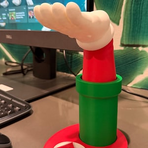 以下が含まれることがあります： 赤、緑、白のデスクアクセサリー。漫画の手のデザインが特徴です。ベースは赤で、白いエンブレム、緑のパイプ、赤い腕が白い手を支えています。このデザインは、人気のあるビデオゲームを連想させます。