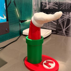 以下が含まれることがあります： 緑色のパイプの形をした3Dプリントのデスクオーガナイザー。赤いベースと、上から伸びる赤と白の腕があります。ベースには赤い「M」ロゴが付いた白い円があります。腕は白で、赤い袖が付いています。