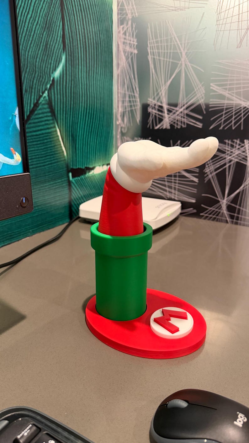 以下が含まれることがあります： 緑色のパイプの形をした3Dプリントのデスクオーガナイザー。赤いベースと、上から伸びる赤と白の腕があります。ベースには赤い「M」ロゴが付いた白い円があります。腕は白で、赤い袖が付いています。