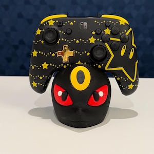 May include: Black Nintendo Switch controller with yellow star accents and a gold cross-shaped button. The controller rests on a black Umbreon-themed stand with red eyes and a yellow ring.