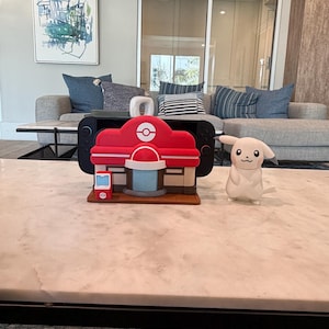 May include: A red and white model of a building with a circular logo, resembling a Pok&eacute;mon Center, sits on a marble table. A white Pikachu figure stands nearby. The building model has a red roof and a blue and white circular structure.
