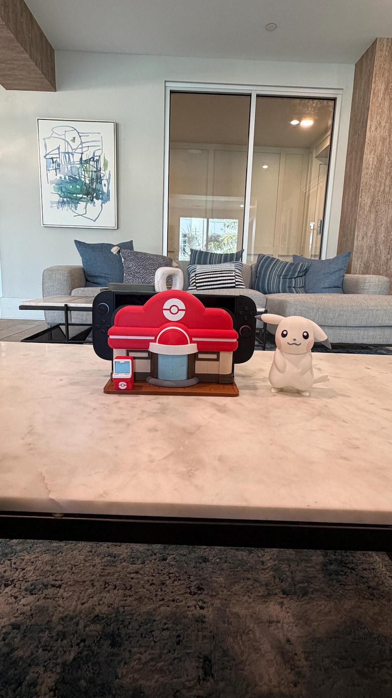 May include: A red and white model of a building with a circular logo, resembling a Pok&eacute;mon Center, sits on a marble table. A white Pikachu figure stands nearby. The building model has a red roof and a blue and white circular structure.