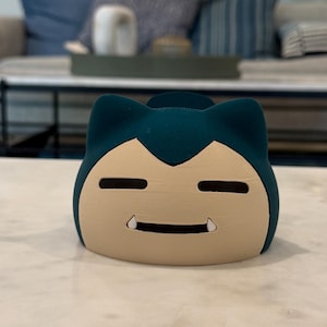 May include: A teal and beige Snorlax-shaped container. The container has a smiling face with two small fangs and two horizontal eye slits. The container is sitting on a white marble surface.