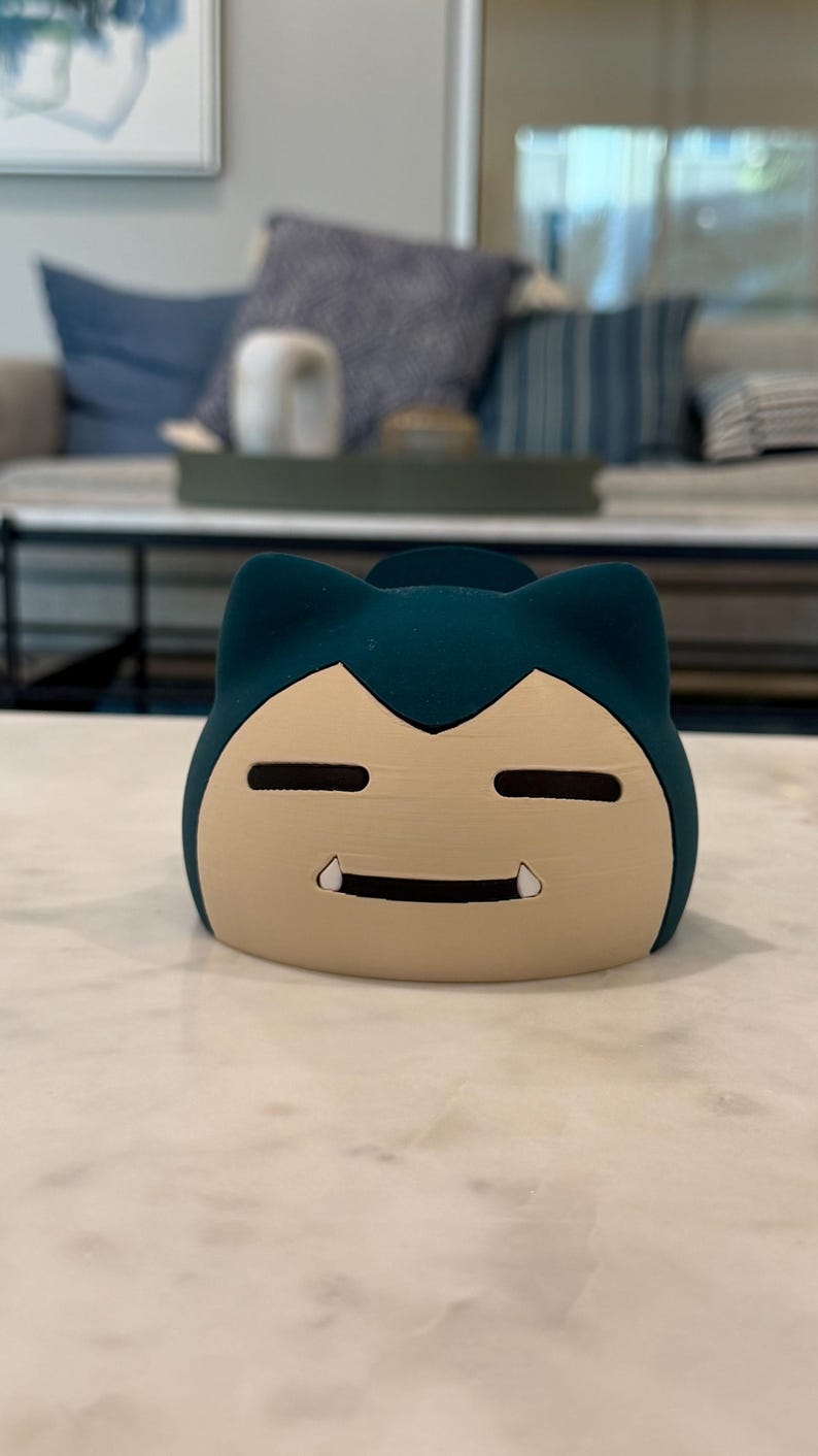 May include: A teal and beige Snorlax-shaped container. The container has a smiling face with two small fangs and two horizontal eye slits. The container is sitting on a white marble surface.