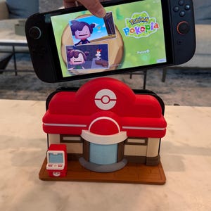 May include: A black Nintendo Switch console displaying the "Pokepia" game, held above a red and white Pokemon Center model. The model features a Poke Ball logo and a small, red vending machine.