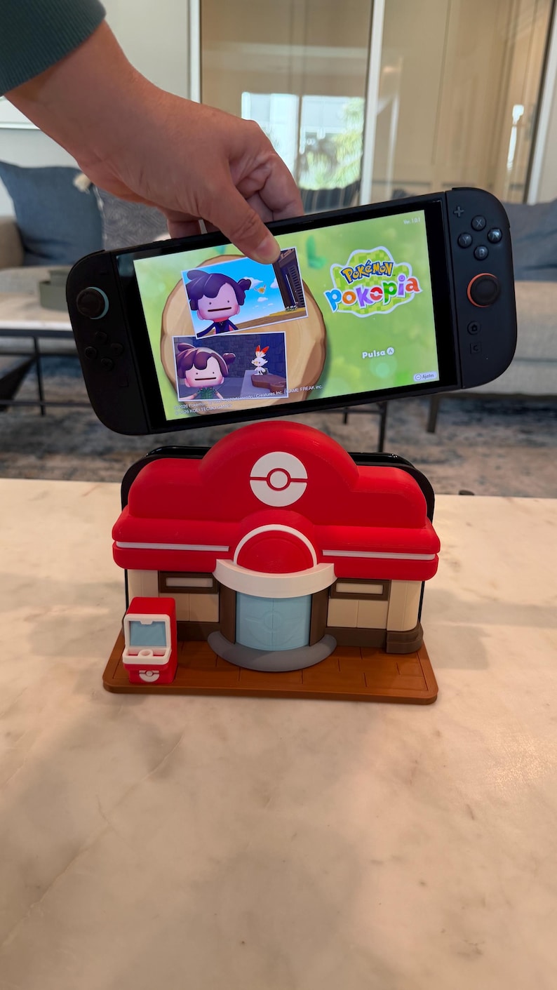 May include: A black Nintendo Switch console displaying the "Pokepia" game, held above a red and white Pokemon Center model. The model features a Poke Ball logo and a small, red vending machine.