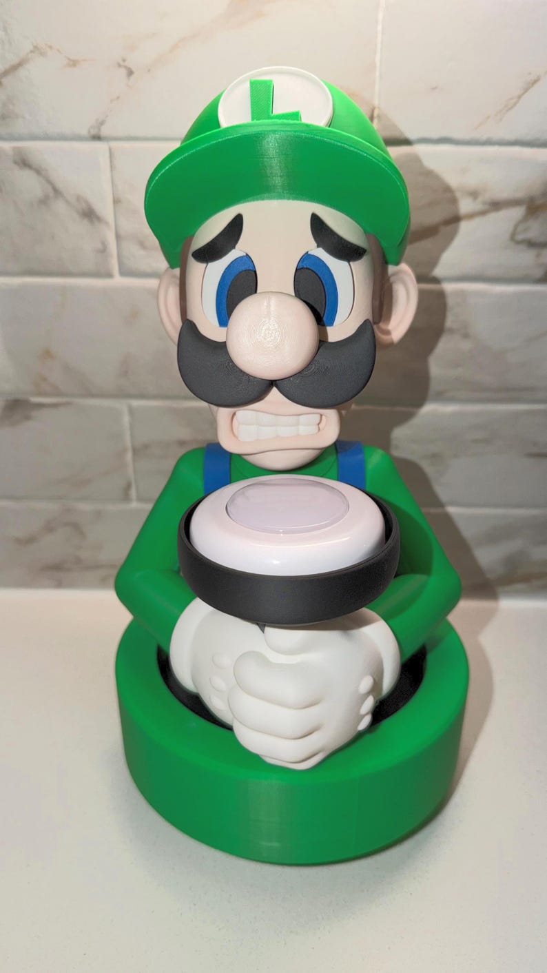 May include: A 3D-printed figurine of Luigi from the Super Mario Bros. series. The green and white figure has a sad expression, a green hat with a white "L", and a black mustache. It holds a white and black circular object.