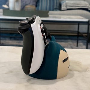 May include: A white and black video game controller on a teal and tan character-shaped stand. The controller has black buttons and a black grip. The stand is in the shape of a cartoon character.