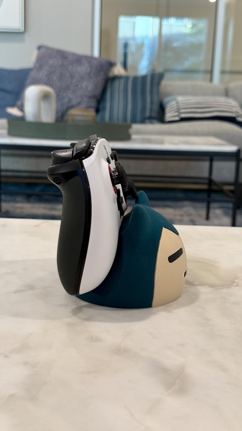 May include: A white and black video game controller on a teal and tan character-shaped stand. The controller has black buttons and a black grip. The stand is in the shape of a cartoon character.