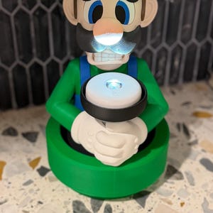 May include: A green and white Luigi figurine with a glowing light. The figurine has a green hat with a white "L" on it, blue overalls, and white gloves. The light is held in the character's hands.