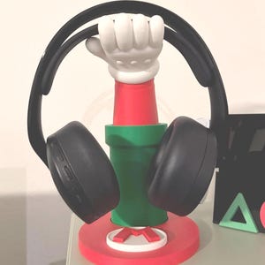 May include: A black headset rests on a red and green headphone stand. The stand features a white gloved hand holding up the headset. The base of the stand is red with a white and red "M" logo.