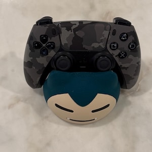 May include: A camouflage-patterned black and grey PlayStation 5 controller rests on a teal and beige Snorlax-shaped stand. The controller features the standard button layout and a central touchpad.