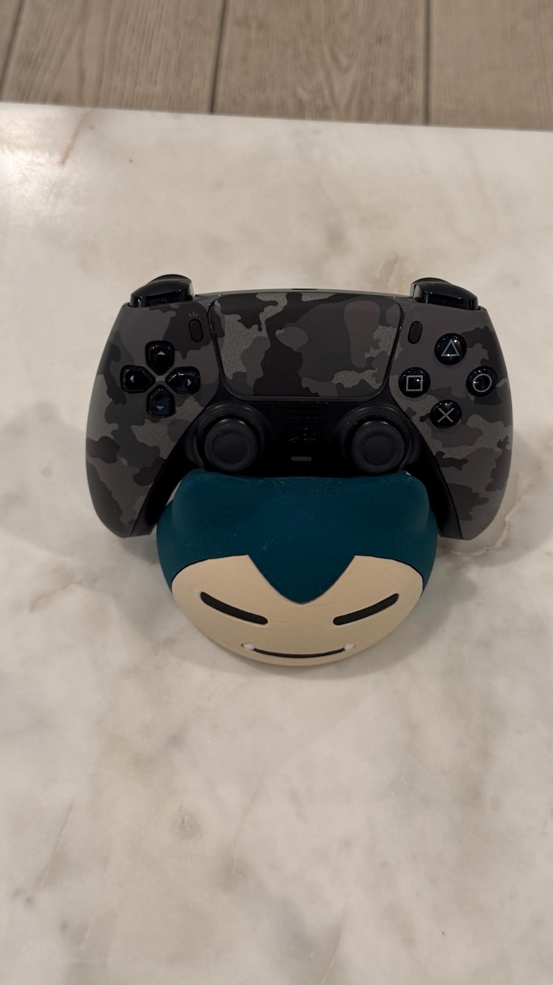 May include: A camouflage-patterned black and grey PlayStation 5 controller rests on a teal and beige Snorlax-shaped stand. The controller features the standard button layout and a central touchpad.