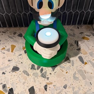 May include: A green Luigi figurine with a glowing green hat and a white circle with a light in the center. The figurine has blue eyes, a black mustache, and a white circle with the letter "L" on the hat.