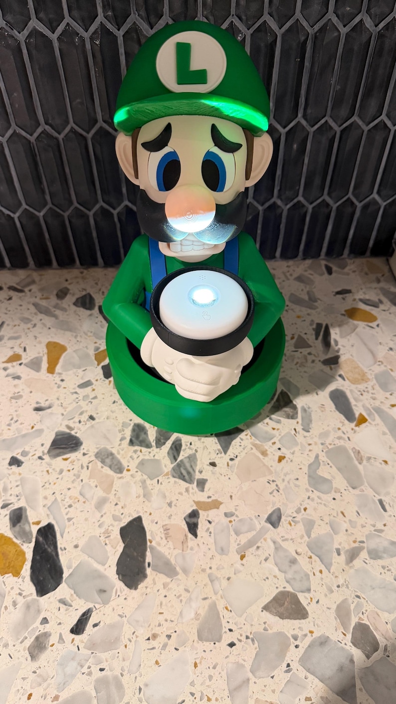 May include: A green Luigi figurine with a glowing green hat and a white circle with a light in the center. The figurine has blue eyes, a black mustache, and a white circle with the letter "L" on the hat.
