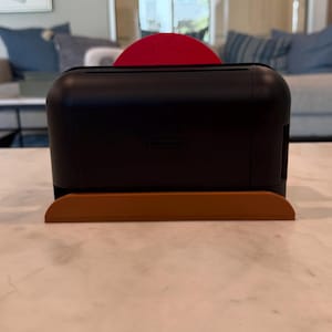 May include: A black Nintendo Switch console with a red Joy-Con controller is placed on a brown stand. The console is rectangular with rounded edges. The stand is a curved, rectangular shape. The Nintendo logo is visible on the console.
