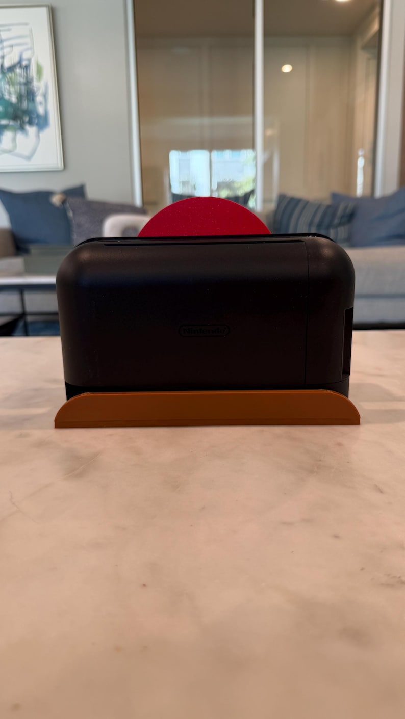 May include: A black Nintendo Switch console with a red Joy-Con controller is placed on a brown stand. The console is rectangular with rounded edges. The stand is a curved, rectangular shape. The Nintendo logo is visible on the console.