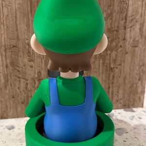 May include: A 3D-printed figurine of a character in a green hat and shirt with blue overalls, standing in a green pipe. The figurine has brown hair and tan skin. The pipe is a bright green color.