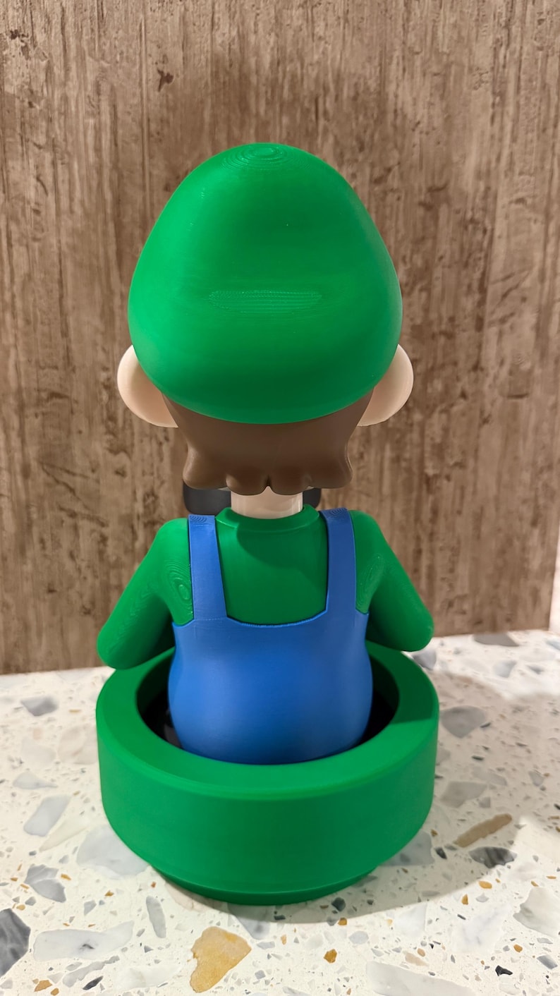 May include: A 3D-printed figurine of a character in a green hat and shirt with blue overalls, standing in a green pipe. The figurine has brown hair and tan skin. The pipe is a bright green color.