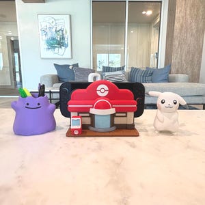 May include: A purple Ditto-shaped pen holder with pens and markers, a red and white Pokemon Center model, and a white Pikachu figure are displayed on a white surface. A black Nintendo Switch is behind the Pokemon Center.