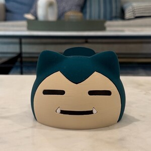 May include: A teal and tan Snorlax-shaped container. The container has a smiling face with small, white teeth. The top of the container is teal, and the face is tan. The container is sitting on a white marble surface.