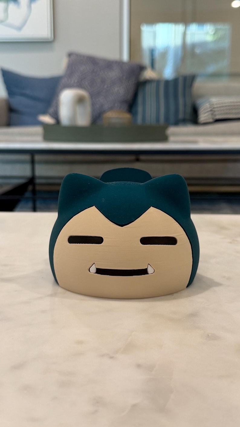 May include: A teal and tan Snorlax-shaped container. The container has a smiling face with small, white teeth. The top of the container is teal, and the face is tan. The container is sitting on a white marble surface.