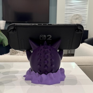 Pode incluir: Um suporte Nintendo Switch em forma de Gengar roxo. O suporte &eacute; um design detalhado impresso em 3D, com um corpo texturizado e uma base em forma de nuvem. Um console Nintendo Switch preto repousa no suporte.