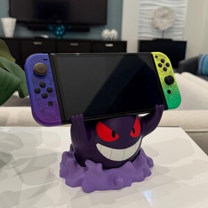 Pode incluir: Um suporte roxo e branco para Nintendo Switch em forma de personagem de desenho animado com olhos vermelhos e um sorriso largo. O suporte suporta um console Switch com controladores roxos e verdes.