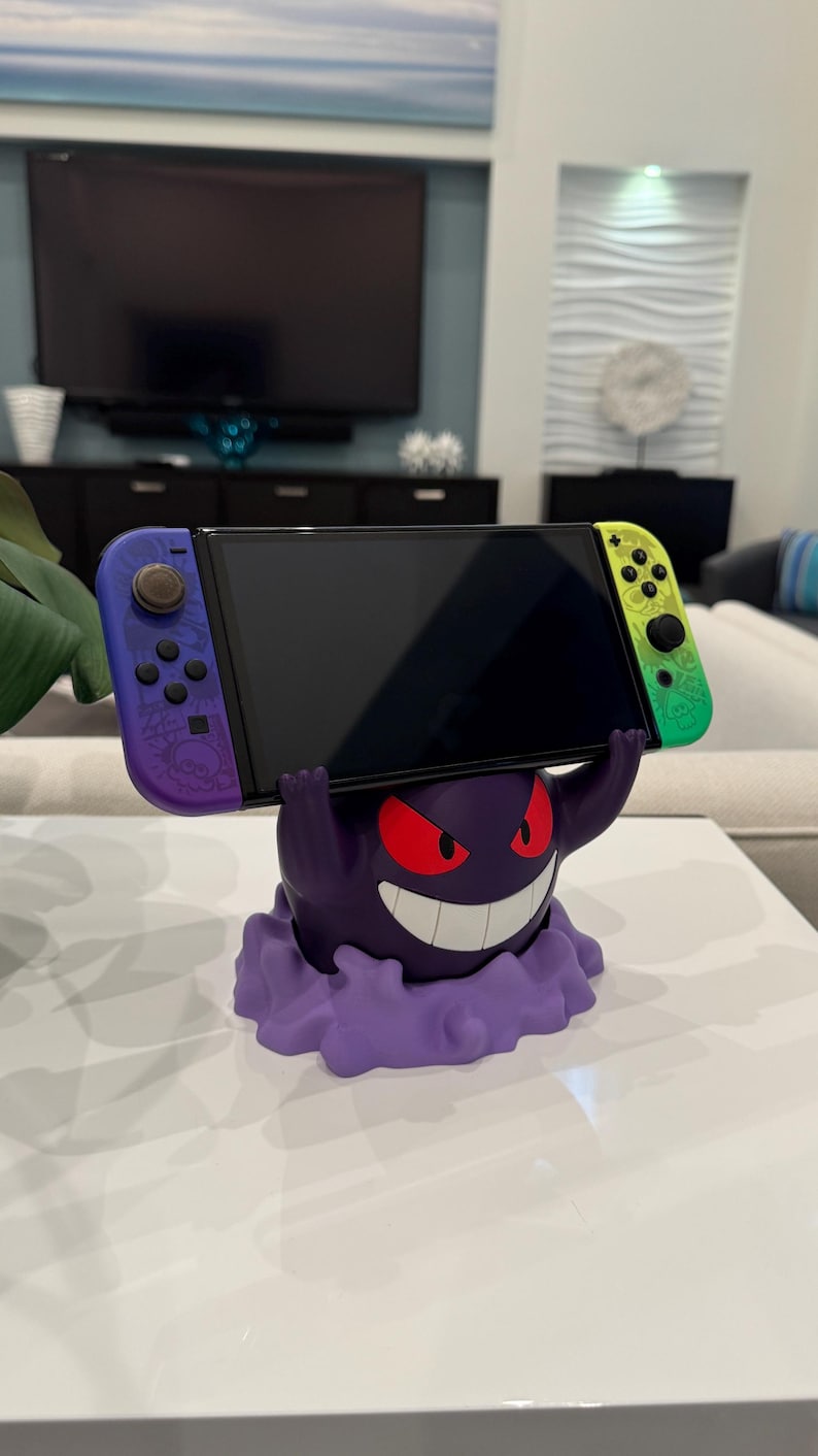 Pode incluir: Um suporte roxo e branco para Nintendo Switch em forma de personagem de desenho animado com olhos vermelhos e um sorriso largo. O suporte suporta um console Switch com controladores roxos e verdes.