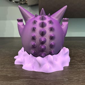 Pode incluir: Uma figura roxa impressa em 3D de um personagem de desenho animado com um corpo redondo, sali&ecirc;ncias pontiagudas e bra&ccedil;os pequenos. A figura est&aacute; em cima de uma base em forma de nuvem, tudo em tons de roxo. &Eacute; um item colecion&aacute;vel.