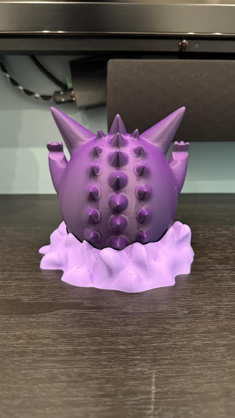 Pode incluir: Uma figura roxa impressa em 3D de um personagem de desenho animado com um corpo redondo, sali&ecirc;ncias pontiagudas e bra&ccedil;os pequenos. A figura est&aacute; em cima de uma base em forma de nuvem, tudo em tons de roxo. &Eacute; um item colecion&aacute;vel.
